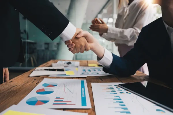 Two business people shaking hands over a desk of financial charts, illustrating a dealership acquisition