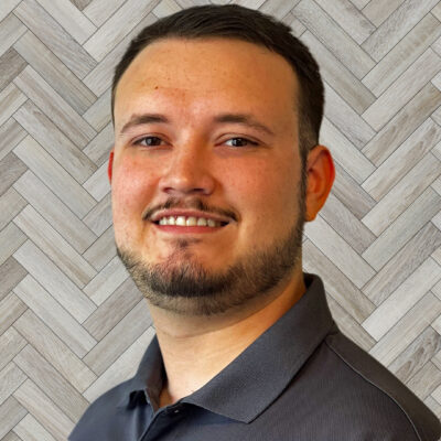 Close-up of a smiling man with stubble beard, wearing a dark polo shirt, against a wood parquet background.